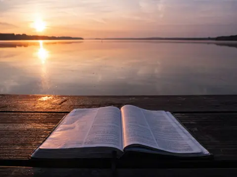 An open book on a wooden dock beside still water at sunrise