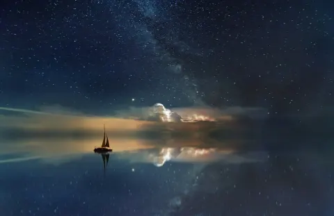 Small sailboat on still water reflecting a star-filled night sky and warm horizon glow